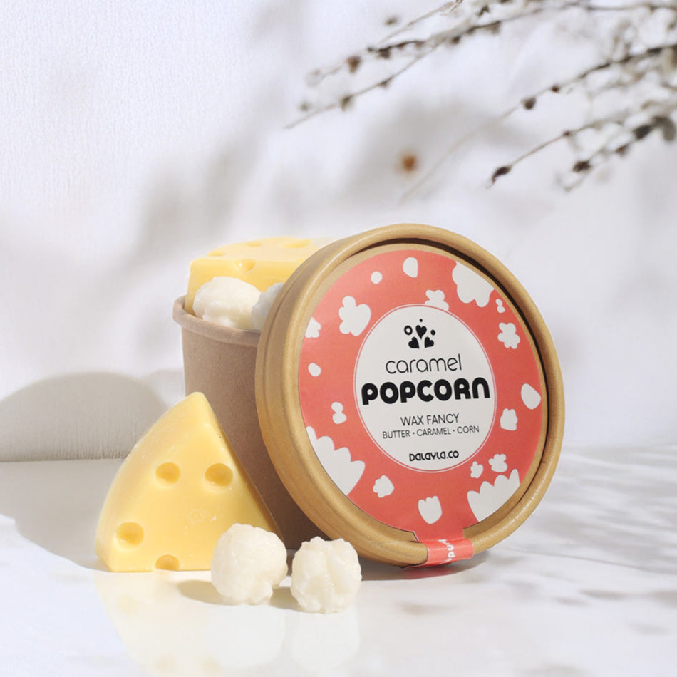 แว็กซ์หอมดีไซน์พิเศษ กลิ่น Caramel Popcorn รูปทรงป๊อปคอร์นและเนยแข็ง วางตกแต่งคู่กับถ้วยคราฟท์ในมุมที่มีแสงธรรมชาติและเงาของกิ่งไม้แบบ Dalayla